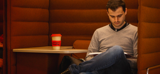A student sits reading in a brown padded pod for studying. There is a desk to his left, with a reusable coffee cup on top of it.