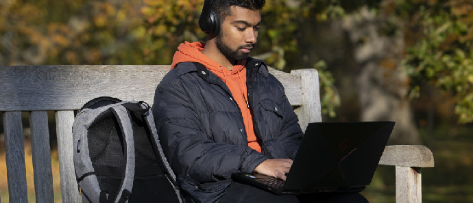 A male student wearing headphones works on his laptop on a bench