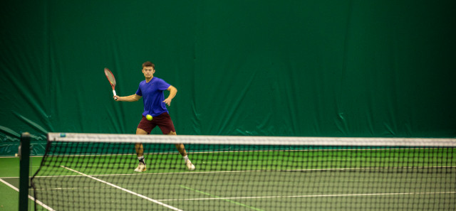A boy in a blue t-shirt is playing tennis inside the Tennis Dome. He has his hand out with his racket about to swing and hit the ball. 
