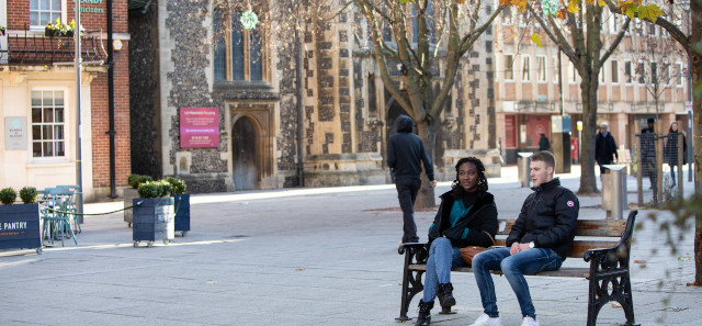 Two students sit on a bench chatting to each other. Behind them is an old church in Reading town centre. 