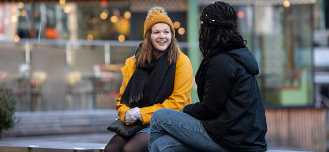 Two students chat to each other. One is wearing a bright yellow coat and is smiling. The other has her back to the camera while while she listens. 
