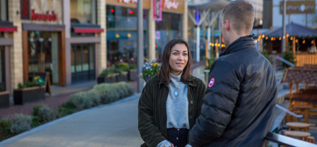 Two students talk to each other while leaning against a rail. Behind them are a row of restaurants. 